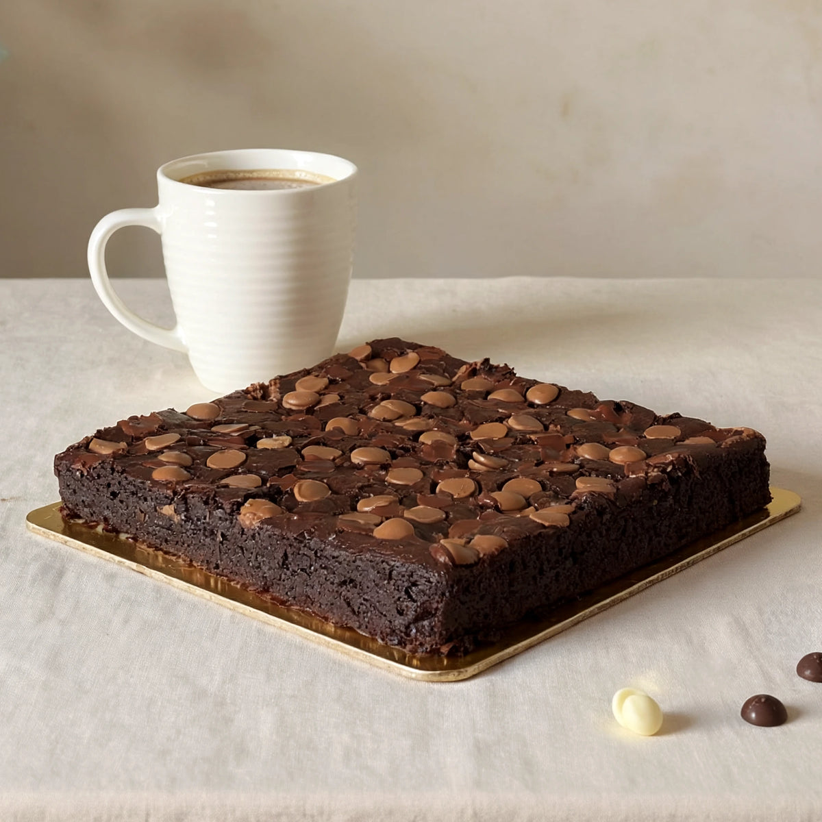 A square, uncut slab of dark chocolate fudgy brownies topped with melted milk chocolate chips on a gold tray, served with a white coffee mug. PuffyClouds Cakes, Kochi
