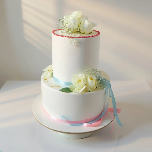 Two-tier elegant floral wedding cake with fresh white roses, delicate baby’s breath, pastel blue and pink ribbons, and gold accents by PuffyClouds, Kochi