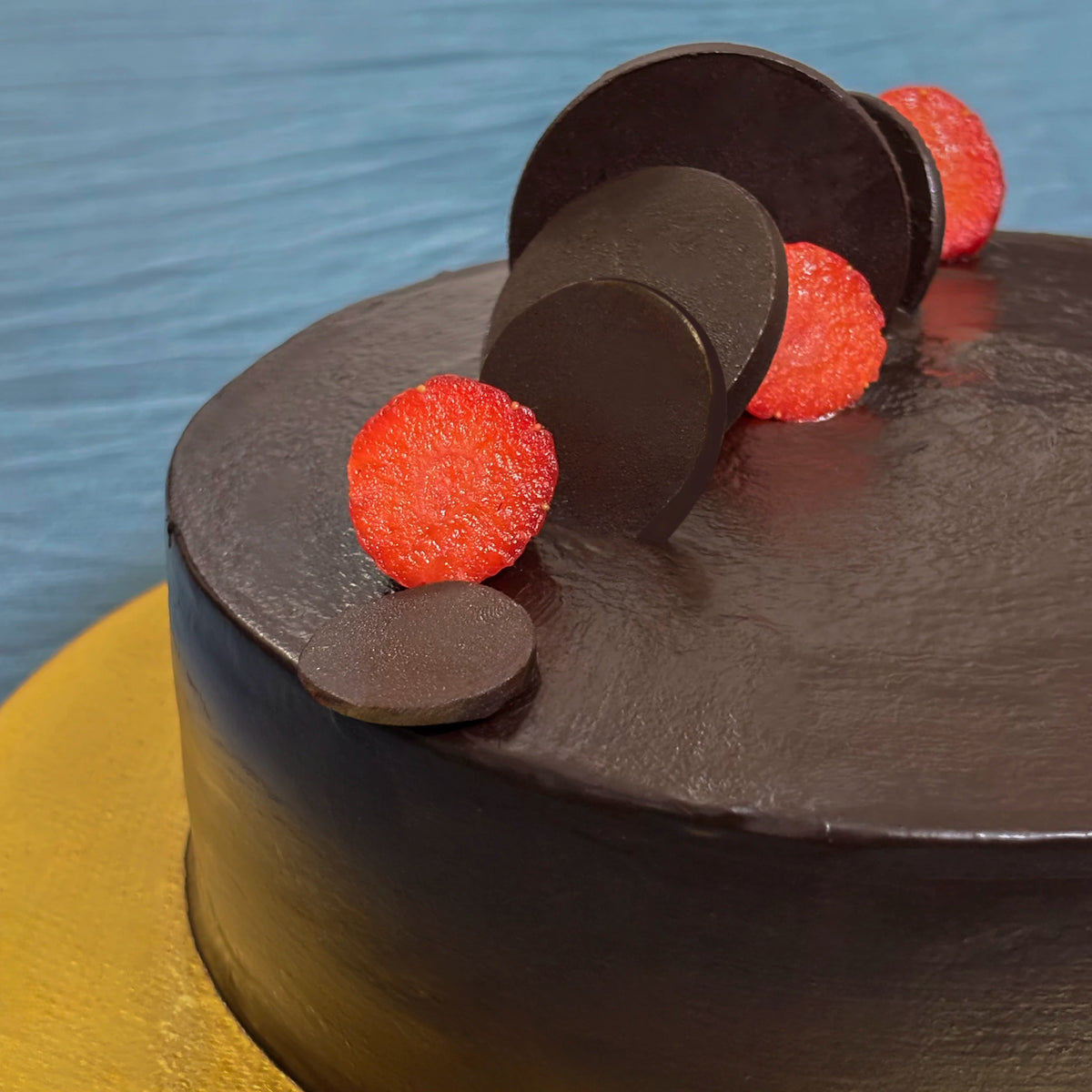 A close-up of a smooth, classic chocolate truffle cake sitting on a textured gold cake board, elegantly decorated with sleek chocolate discs and fresh strawberry slices.