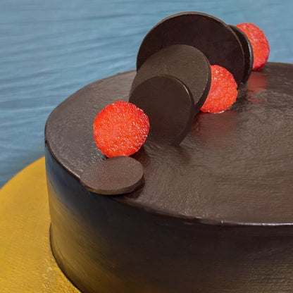 A close-up of a smooth, classic chocolate truffle cake sitting on a textured gold cake board, elegantly decorated with sleek chocolate discs and fresh strawberry slices.