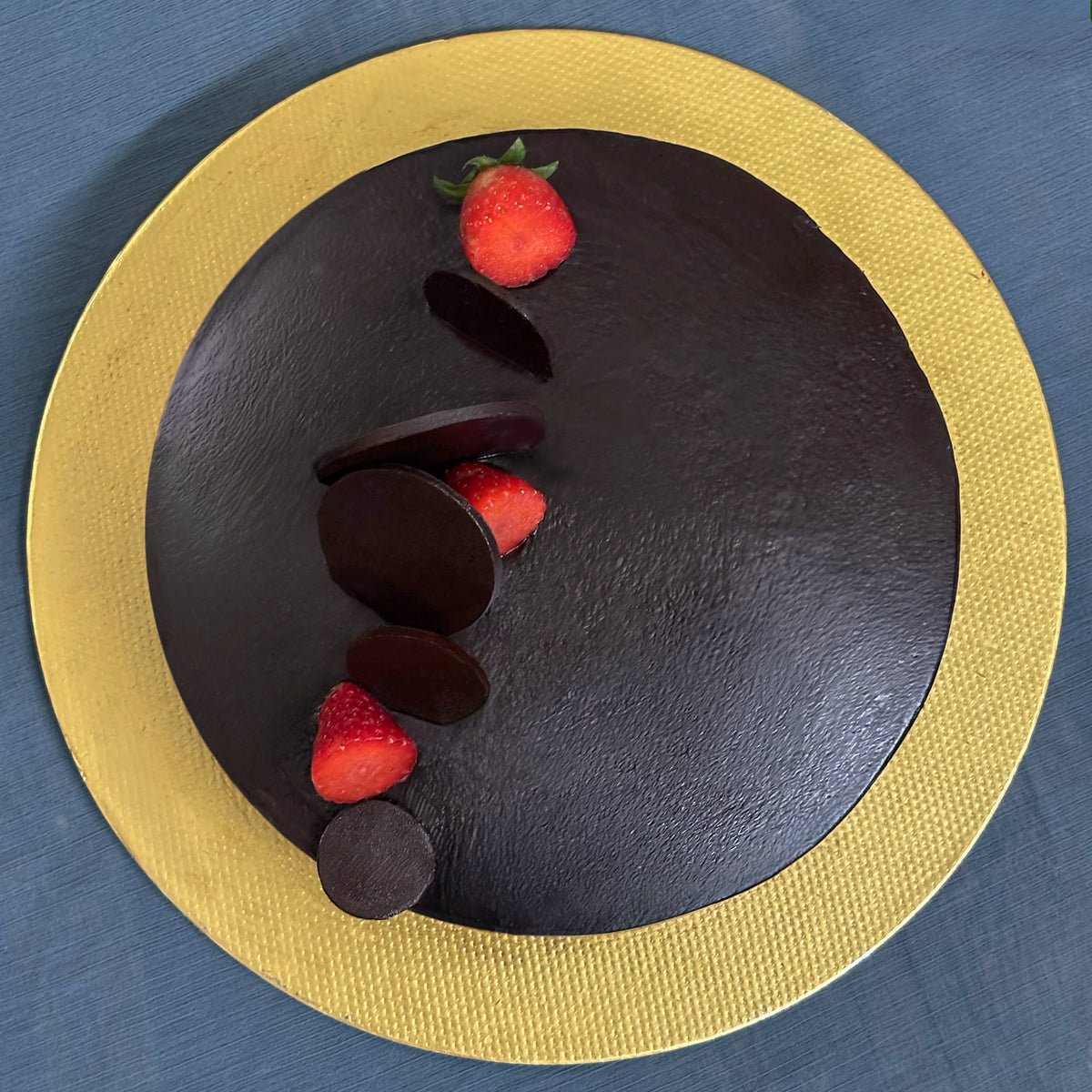 A round classic chocolate cake topped with strawberries and chocolate slices, presented on a gold  cakebase against a blue textured background.