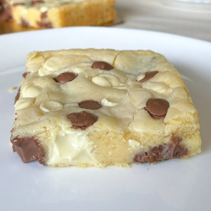 A thick Kinder blondie bars on a white plate, showcasing a soft, gooey texture filled with melted white and milk chocolate pieces. From PuffyClouds, Kochi
