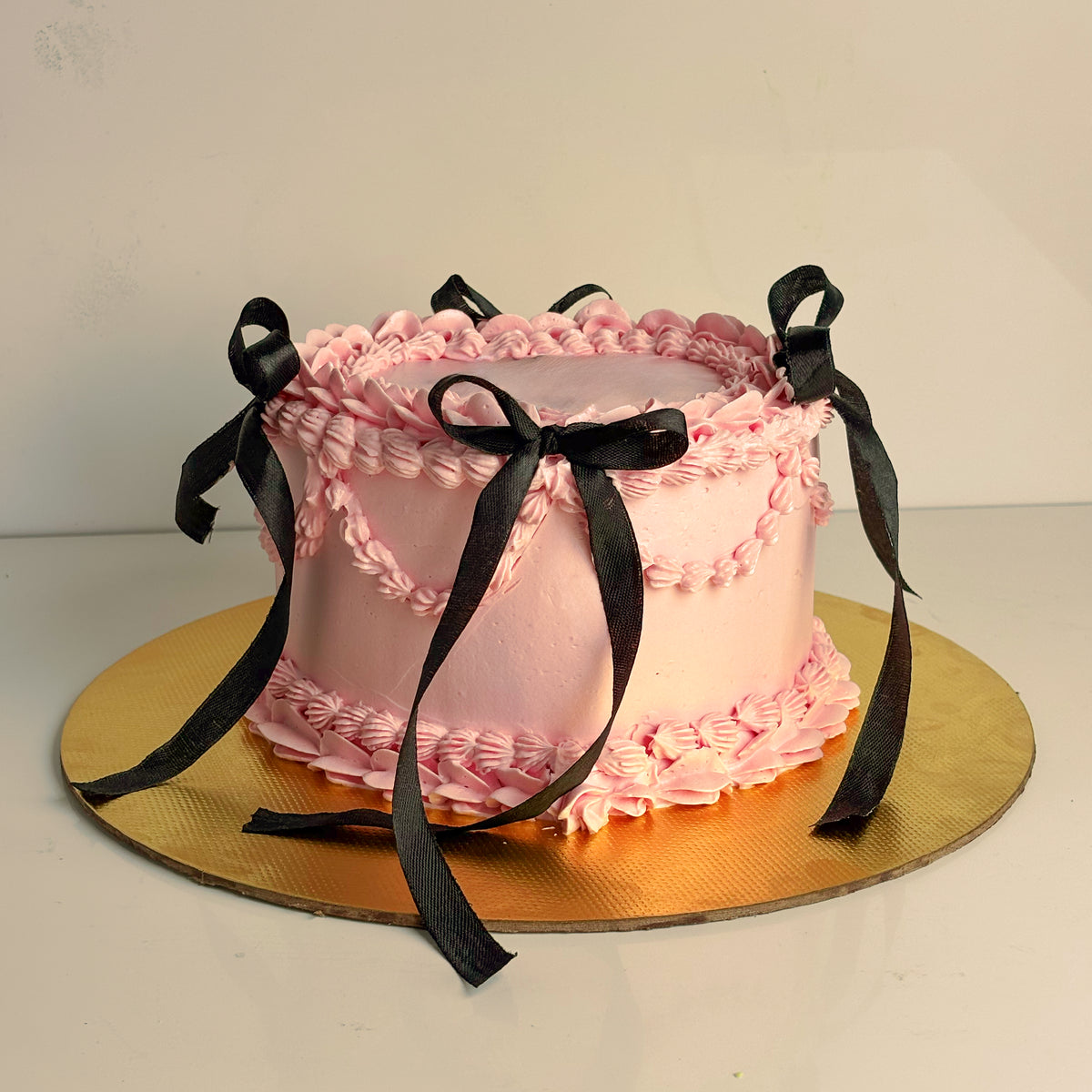 Premium pink vintage Lambeth buttercream celebration cake decorated with hand-tied black satin ribbon bows on a gold cake board by PuffyClouds Kochi.
