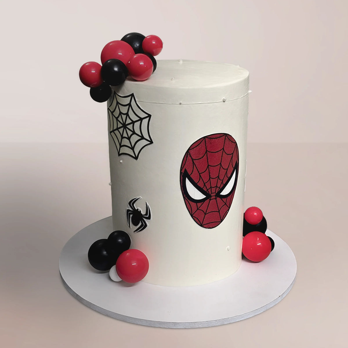 A tall, smooth white buttercream Spider-Man theme cake decorated with a red fondant mask cutout, black spider webs, and glossy red and black chocolate sphere clusters, handcrafted by PuffyClouds in Kochi.