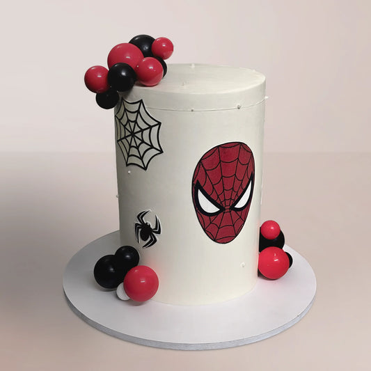 A tall, smooth white buttercream Spider-Man theme cake decorated with a red fondant mask cutout, black spider webs, and glossy red and black chocolate sphere clusters, handcrafted by PuffyClouds in Kochi.